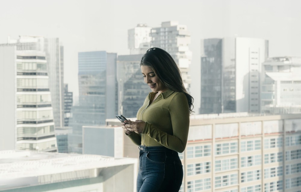 Mulher usando um celular diante das janelas de um edifício, com a cidade ao fundo.