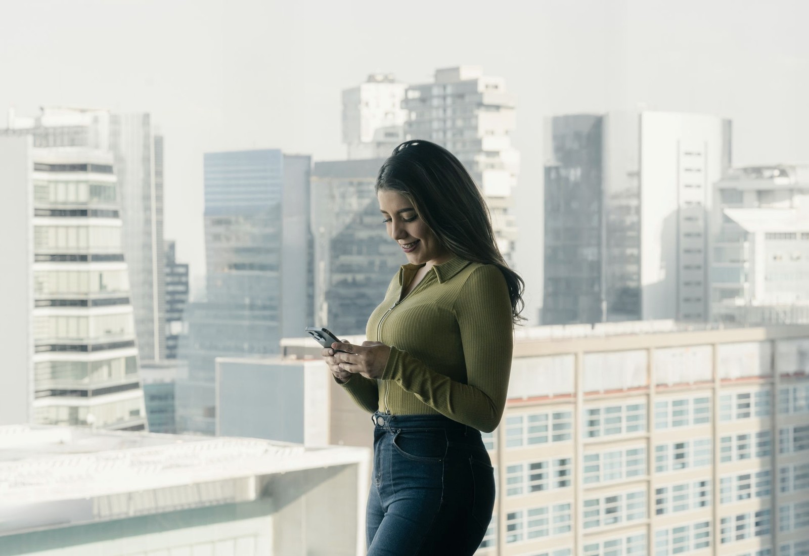 Mulher usando um celular diante das janelas de um edifício, com a cidade ao fundo.